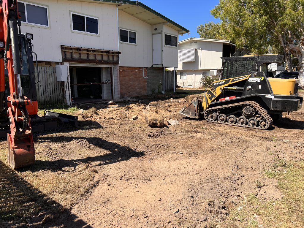 Excavator and tracked loader clearing a yard beside a raised house